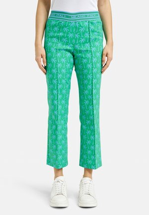 Grüne Cropped-Hosen mit einem hellblauen Palmbaum-Muster, ausgestattet mit einem breiten Bündchen, das "MILANO ITALY" trägt. Getragen mit weißen Sneakers.