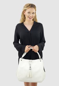 White leather handbag with a textured finish. Features silver hardware, dual handles, and a matching tassel. Displayed by a person in a black dress.