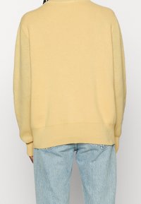 Person klädd i en lös, blekgul tröja och ljusblå denimjeans, sedd bakifrån mot en enkel bakgrund.