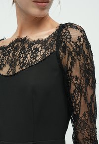 Robe noire avec un corsage ajusté orné d'une superposition en dentelle sur les épaules, des motifs floraux complexes et des embellissements en perles le long de l'encolure.