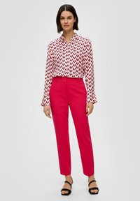 Chemise blanche à boutons ornée de flèches noires et rouges, associée à un pantalon ajusté rose vif et des sandales noires à lanières.