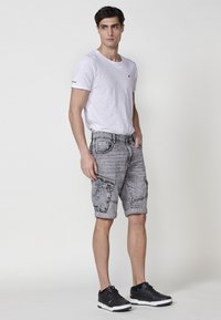 Shorts en denim gris avec poches cargo et ourlets roulés, associés à un t-shirt blanc à col rond et des baskets noires. Design décontracté, coupe relaxée.