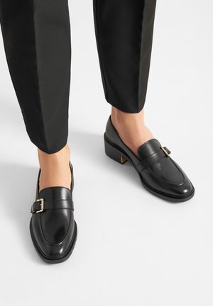 Mocasines bajos de cuero negro con detalle de hebilla dorada, usados con pantalones negros cortos y pies descalzos.