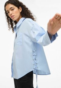 Chemise oversize bleu clair en tissu lisse, dotée d'une poche avant et d'un détail unique à lacets sur le côté avec des œillets métalliques.