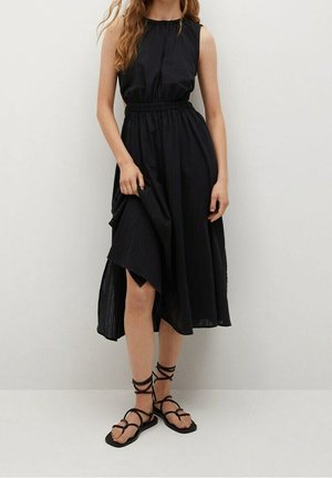 Vrouw draagt een zwarte mouwloze midi-jurk met een geplooide taille, houdt de rok vast en zwarte platte vetersandalen tegen een effen witte achtergrond.