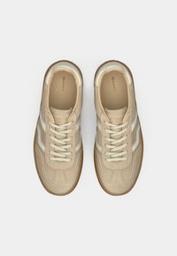Μπεζ αθλητικά παπούτσια από σουέτ και δέρμα, με δύο λευκές ρίγες, στρογγυλή μύτη και καφέ καουτσούκ εξωτερική σόλα με υφή. Κορδόνια στο ίδιο μπεζ χρώμα.