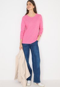 Un haut rose à manches longues en tissu doux et texturé, associé à un jean bleu à jambes larges. Des baskets blanches et une veste beige sont drapées sur un bras.