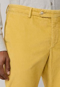 Keltaiset corduroy-housut pystyribatuilla kuvioilla, edessä napitus ja sivutaskut, esitelty yhdessä vaalean kuvioidun paidan kanssa.