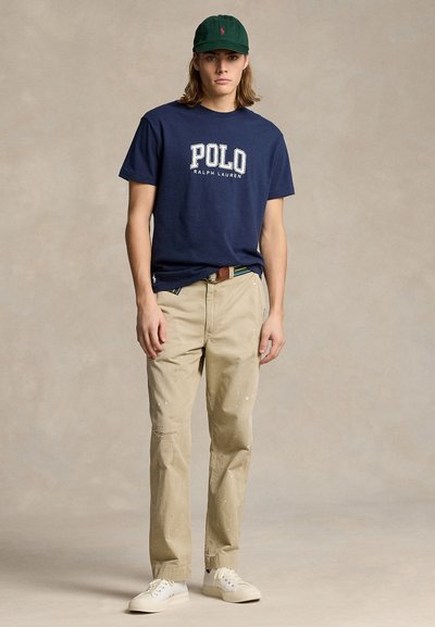 Young man wearing a navy Polo Ralph Lauren t-shirt, beige pants with paint splatters, white sneakers, and a dark green baseball cap, standing.