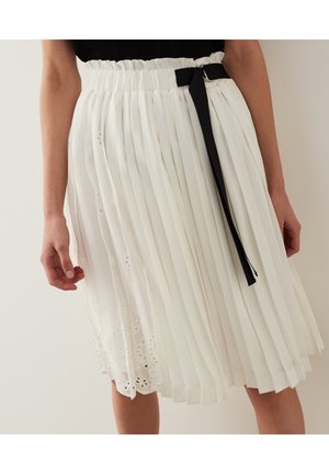 Femme portant une jupe plissée blanche avec des détails en dentelle œillet et une ceinture en ruban noir, associée à un haut noir.
