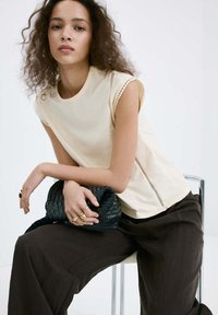 Jeune femme aux cheveux bouclés assise sur une chaise, portant un haut beige sans manches, un pantalon large foncé, tenant une pochette noire tressée avec des anneaux dorés.