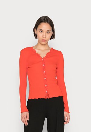 Camisola longa de manga comprida vermelha com decote ondulado e acabamento de renda decorativa. Apresenta fecho de botão na parte da frente e punhos das mangas rufados.