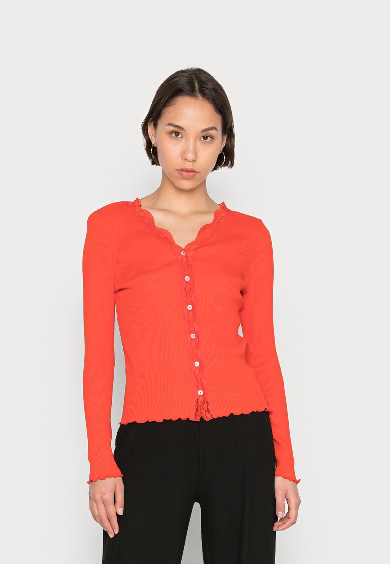 Camisola longa de manga comprida vermelha com decote ondulado e acabamento de renda decorativa. Apresenta fecho de botão na parte da frente e punhos das mangas rufados.