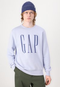 Ανοιχτό μωβ φούτερ με τη λέξη "GAP" σε σκούρο ναυτικό μπλε γράμματα, στρογγυλή λαιμόκοψη, και ριμπά μανίκια και στρίφωμα. Συνδυάζεται με ελιά πράσινα παντελόνια και μπλε σκουφί.