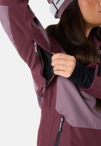 Giacca bordeaux e lilla chiaro con zip di ventilazione. Presenta una fodera interna nera e polsini regolabili. Design elegante e funzionale per il layering.