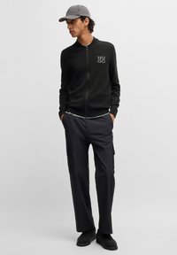 Suéter con cremallera en verde oscuro con el logotipo "HUGO", combinado con pantalones cargo negros que tienen bolsillos laterales. El modelo lleva una gorra gris y zapatos negros.