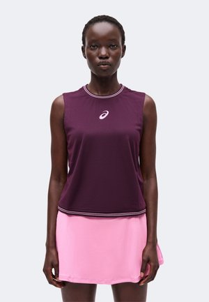 Femme portant un haut de sport sans manches violet foncé et une jupe rose clair, debout devant un fond clair uni.