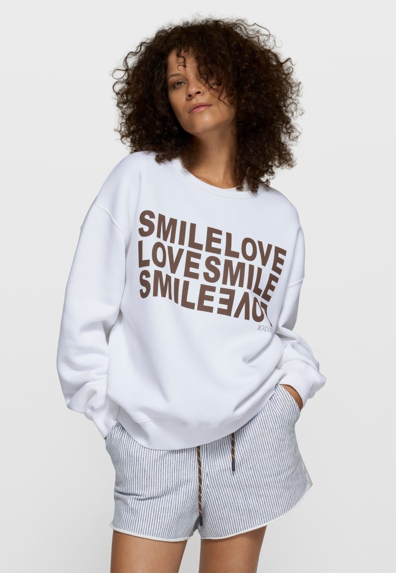 Sudadera blanca oversize con texto gráfico marrón: "SMILE LOVE" repetido. Combinada con pantalones cortos a rayas azules y blancas.
