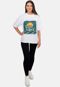 Wit katoenen t-shirt met een grafische print van een zon en golven met de tekst "Good Vibes." Gecombineerd met zwarte legging en witte sneakers.