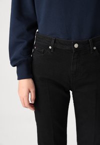Black denim jeans with straight cut, five-pocket design, silver buttons, and visible stitching. Worn with a navy sweatshirt.