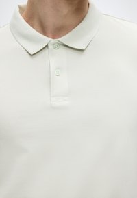 Φωτεινό μπεζ πόλο μπλουζάκι με υφασμάτινη υφή· διαθέτει κλασικό γιακά, δύο κουμπιά και μινιμαλιστικό σχέδιο.