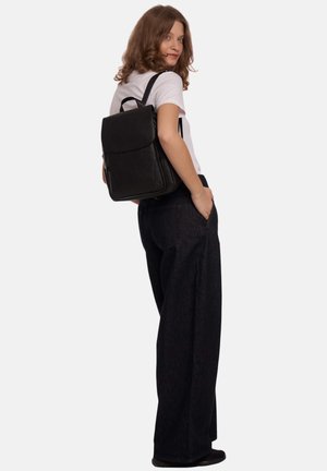 Jonge vrouw draagt een wit overhemd en zwarte wijde broek, met een zwarte leren rugzak over haar schouder.