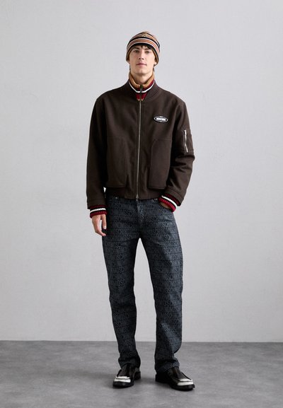 Brun zip-up jacka med ribbad röd och vit mudd, kombinerad med svarta mönstrade byxor med "Moschino" text. Svarta skor och en randig mössa.