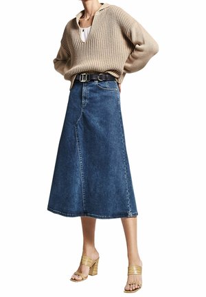 Femme portant un pull en tricot beige, une jupe midi en denim bleu avec une ceinture noire, et des sandales à talons beiges, se tenant devant un fond blanc.