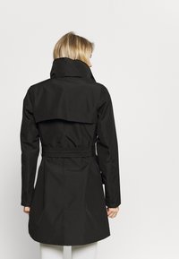 Cappotto nero con colletto alto, vita regolabile con cintura e fondo svasato. Presenta una texture liscia e opaca e un design semplice.
