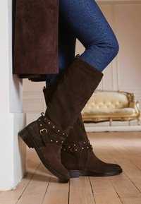 Hnědé semišové kozačky po kolena se zlatými hroty a pásky. Mají boční zip a nízký podpatek, umístěné na pozadí z dřevěné podlahy.