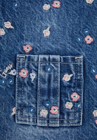 Blue denim fabric featuring a small, embroidered pocket with floral patterns in pink, white, and blue. Stitched logo reads "Guess."