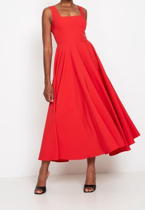 Kobieta w czerwonej sukience midi bez rękawów z kwadratowym dekoltem i czarnymi sandałami na obcasie z odkrytymi palcami, stojąca na tle białego tła.