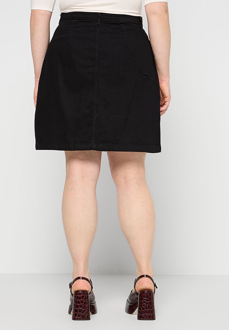 Schwarzer Jeansrock mit geradem Schnitt, sichtbarer Steppnaht und Bündchen. Das Model trägt braune Schuhe mit Krokodilprägung und Absatz.