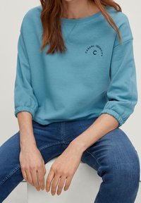 Sweatshirt bleu clair avec un col rond, des poignets élastiques et une coupe décontractée. Présente le texte "IDENTITÉ DÉCONTRACTÉE" brodé à l'avant.