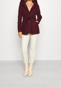 Burgunderröd huvjacka med knyt i midjan, två framfickor och knappstängning, kombinerad med krämfärgade figurnära byxor och beige ankelstövlar.