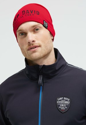 Man wearing red beanie with "Camp David" text and navy jacket with "Camp David Sail" patch, facing slightly left on plain background.