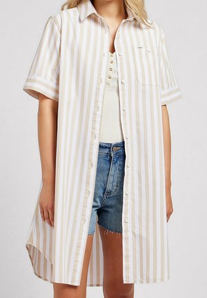 Vrouw draagt een beige-wit gestreepte jurk met korte mouwen en knopen, over een wit geribd topje en blauwe spijkershorts.