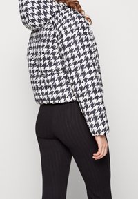Kvinna klädd i svart och vit houndstooth puffjacka med huva och svarta ribbade leggings, visad bakifrån mot en enfärgad bakgrund.