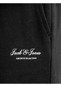 Tissu noir avec le logo cursif blanc "Jack & Jones" au-dessus du texte "ARCHIVE SELECTION", montrant une couture et une partie d'une poche.