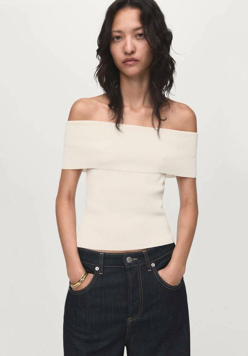 Romantische off-the-shoulder crèmekleurige top met een getailleerde pasvorm, gecombineerd met donkerdenim hoge taille jeans met zakken en een gouden accentarmband.