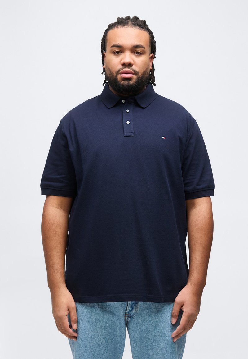 Άνδρας με πλεξούδες στα μαλλιά και μούσι, φορώντας μπλε πόλο πουκάμισο και ανοιχτόχρωμο τζιν, στέκεται μπροστά σε απλό λευκό φόντο.