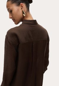 Bruine, lichte shirt met kraag en lange mouwen, voorzien van een subtiele textuur en een rugpanddetail. Gouden druppeloorbellen zichtbaar.