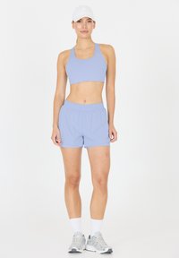 Vaaleansininen urheilbrassi ja siihen sopivat shortsit, jotka ovat pehmeää materiaalia. Mallilla on päässään valkoinen lippis ja harmaat urheilukengät valkoisten sukkien kanssa.