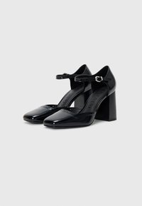 Schwarze Lacklederschuhe mit spitzem Zehenbereich, Knöchelriemen und strukturiertem Akzent. Der Blockabsatz misst etwa 8 cm.