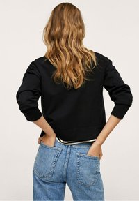 Femme aux cheveux longs et ondulés portant un sweat-shirt noir et un jean bleu, les mains dans les poches arrière, debout devant un fond uni.