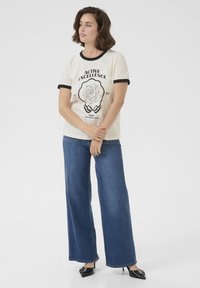 T-shirt crème avec un col et des manches noires, présentant une impression graphique noire d'une rose, associé à un jean bleu ample et des talons noirs.