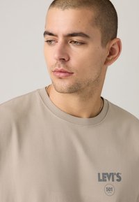 T-shirt de algodão bege com gola redonda e logotipo da Levi's impresso em cinza no lado esquerdo do peito. Textura lisa.