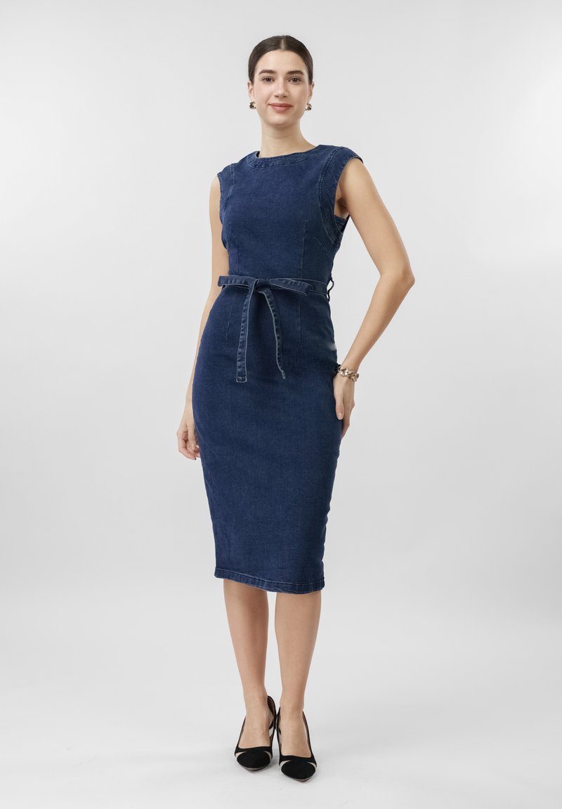 Γυναίκα που στέκεται φορώντας στενή, αμάνικη, μεσαίου μήκους, σκουρόχρωμη μπλε τζιν φόρεμα με ζώνη και μαύρες μυτερές γόβες, πάνω σε απλό φόντο.