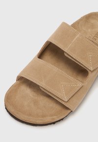 Beige velúr szandál két állítható pánttal, nyitott kialakítással és lapos talppal. Jellemzője a steppelt díszítés és a sima bélés.
