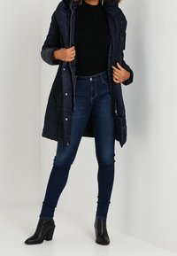 Navy puffermantel med dragkedja och tryckknappar, bärs över en svart tröja. Matchas med mörkblå skinny jeans och svarta klackstövlar.